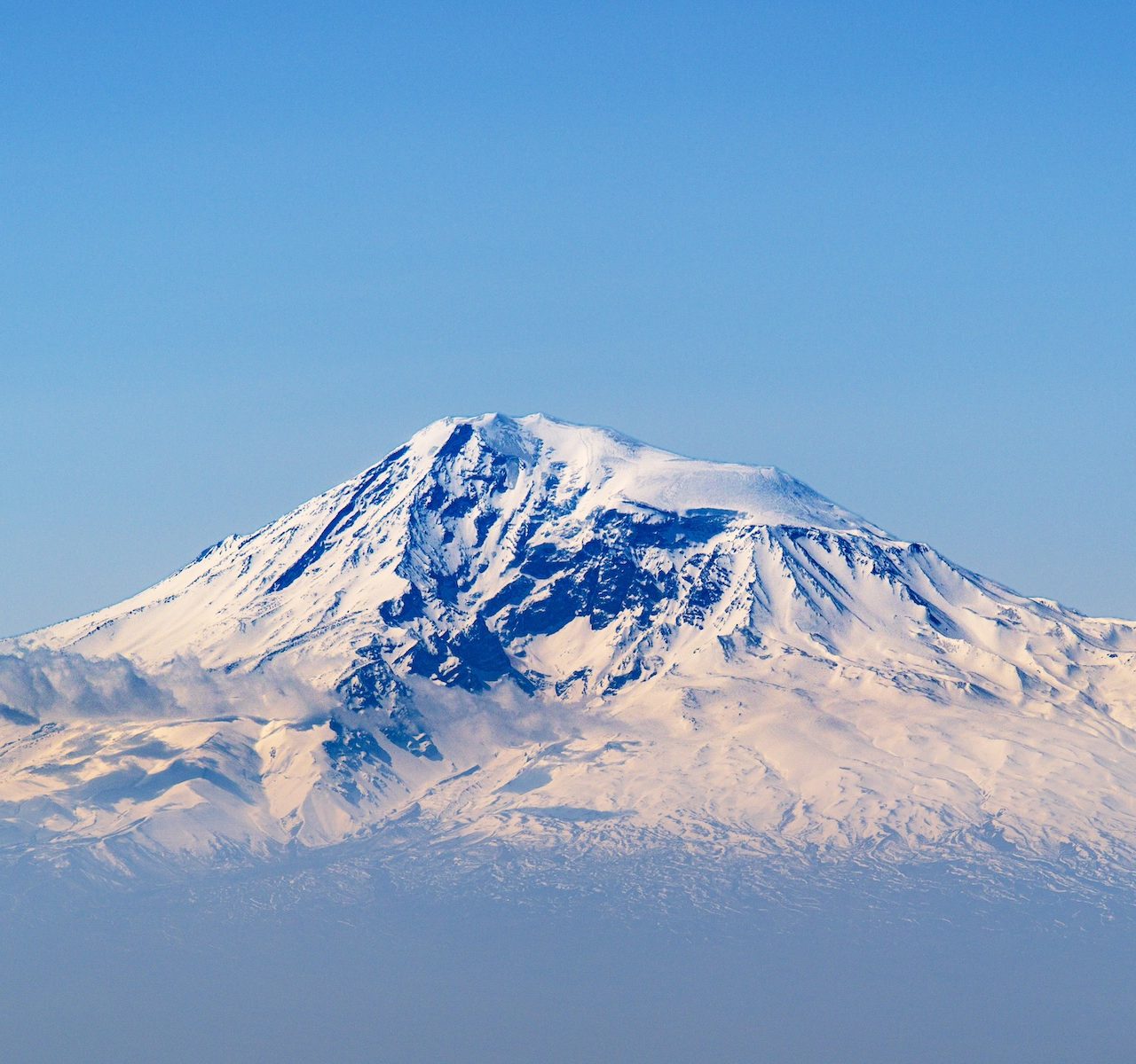 Snow-capped mountain rises above misty clouds. Clear blue sky surrounds peak